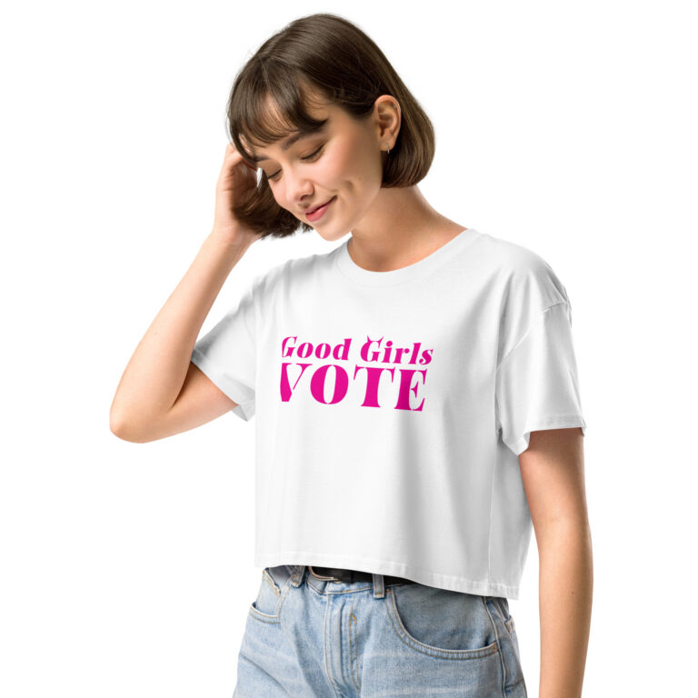 Young woman wearing a white crop tee with bratty pink Good Girls Vote stencil lettering, tucked into light jeans with a black belt